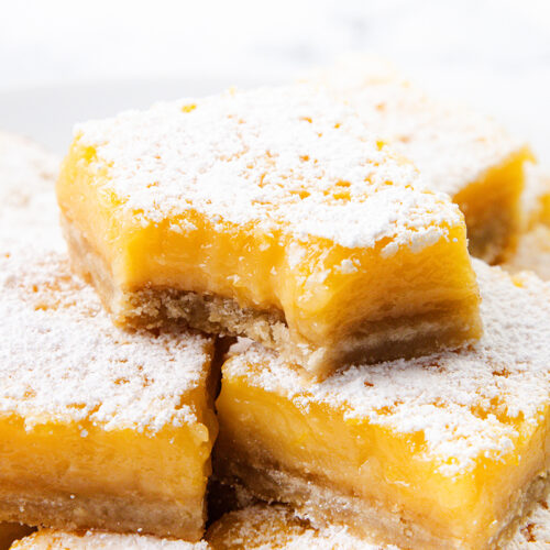 A close-up of lemon bars stacked on a plate, topped with powdered sugar. One bar is on top with a bite taken out, revealing the creamy filling and thick shortbread crust—just like the Best Lemon Squares Recipe.