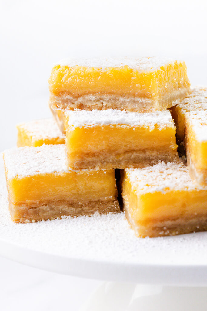 A close-up of stacked lemon bars made with the Best Lemon Squares Recipe, featuring a vibrant yellow filling and powdered sugar topping, displayed on a white surface.