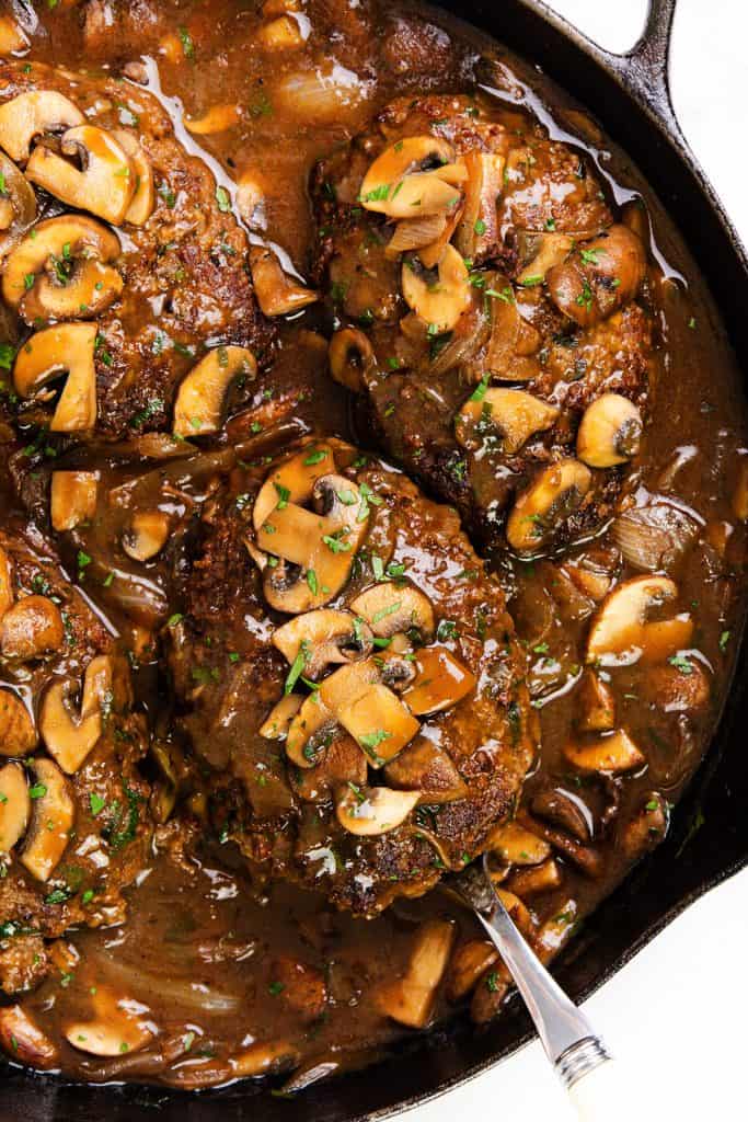 A skillet filled with Salisbury Steak patties, covered in rich brown mushroom gravy and garnished with sliced mushrooms and chopped herbs. A silver spoon rests under one Salisbury Steak patty.