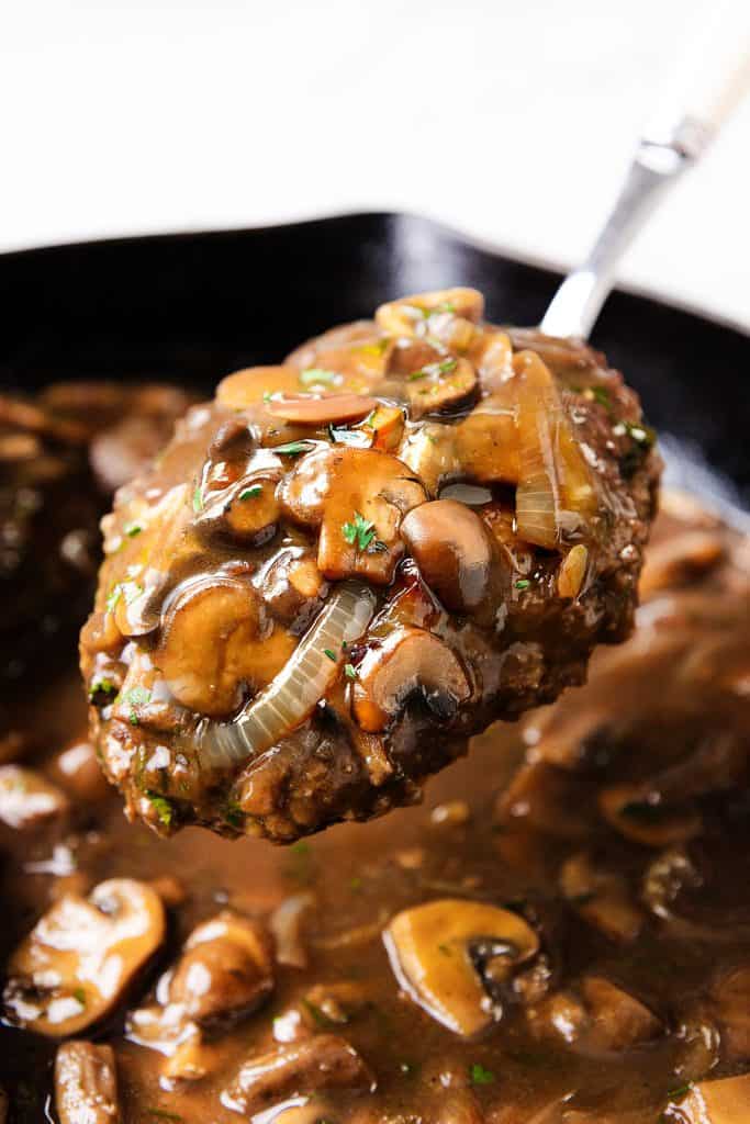 A close-up of a spoon lifting Salisbury Steak topped with sautéed mushrooms, onions, and rich brown gravy from a skillet filled with more steak, mushrooms, and savory sauce.