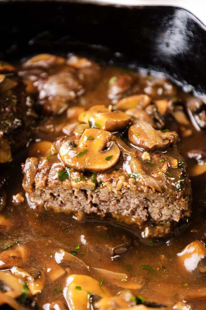 Close-up of a Salisbury Steak topped with sliced mushrooms and rich brown gravy, served in a cast iron skillet. The steak is cut open to reveal its juicy interior.