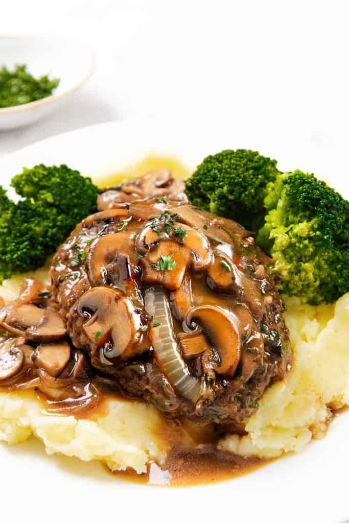A plate of creamy mashed potatoes topped with juicy Salisbury Steak, smothered in mushroom and onion gravy, garnished with parsley, and served alongside fresh steamed broccoli.