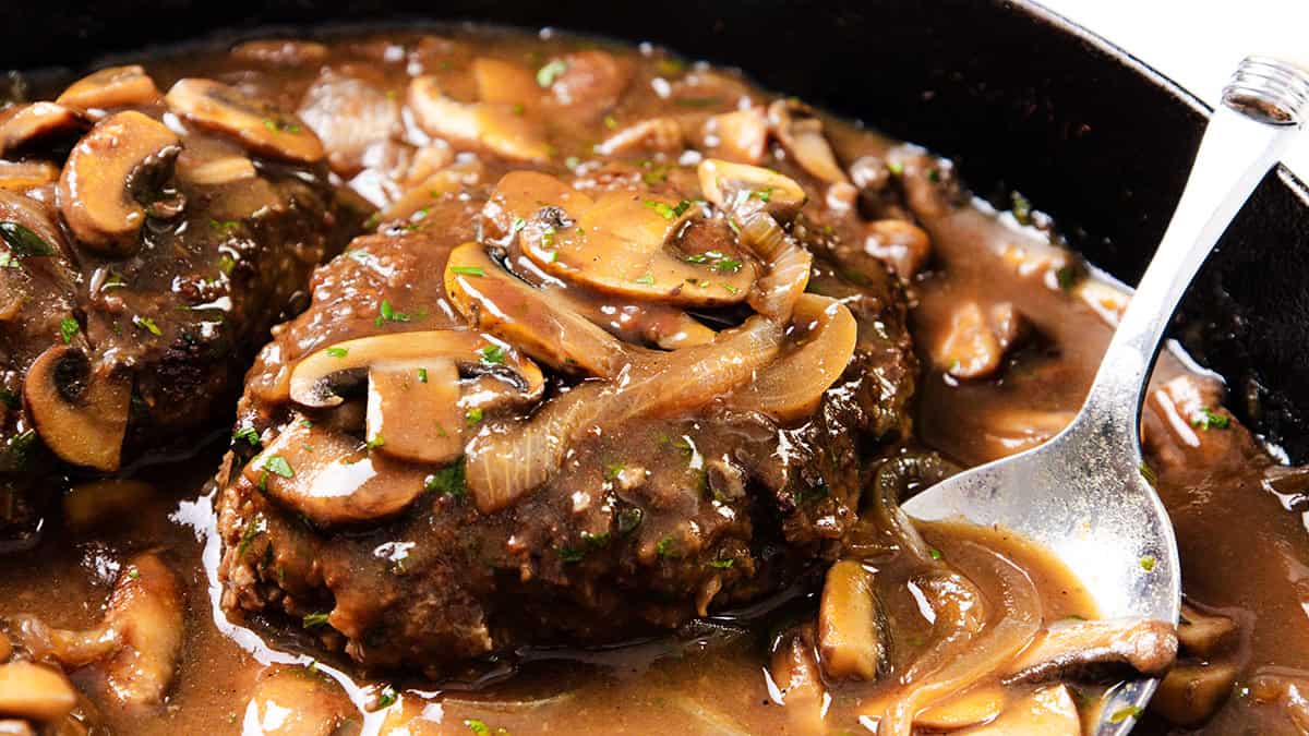 A close-up of Salisbury Steak covered in savory brown mushroom and onion gravy, with sliced mushrooms and onions visible. A spoon rests in the rich sauce in a dark skillet.