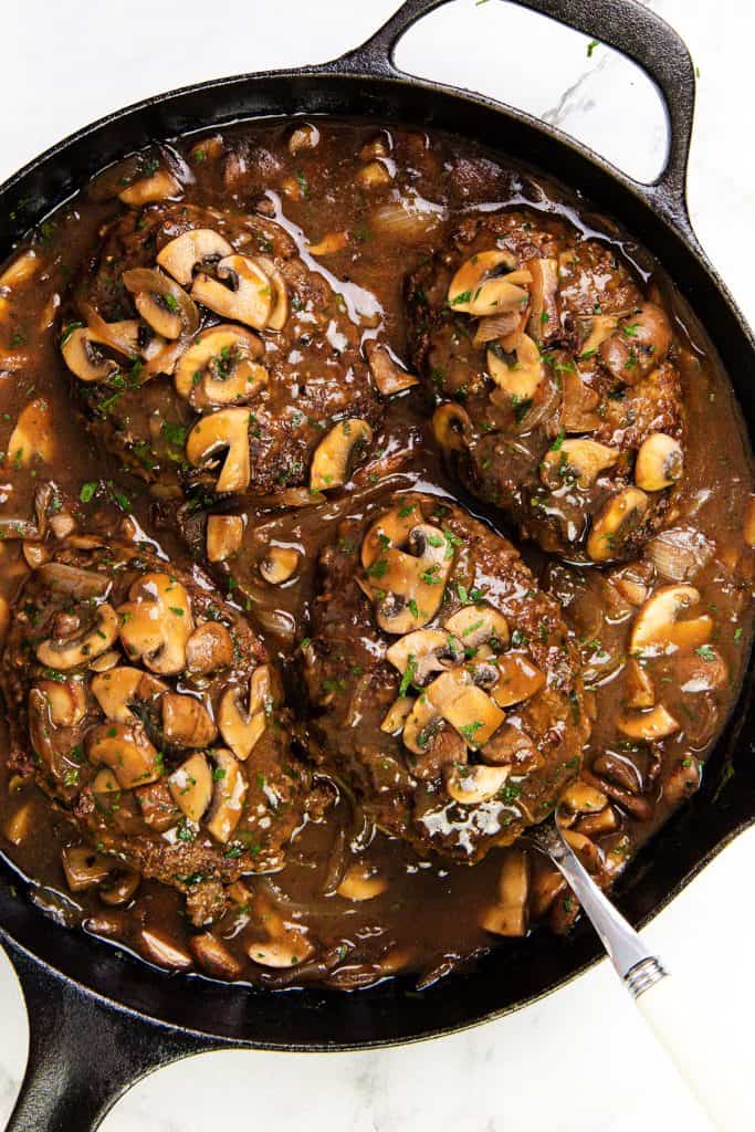 4 Salisbury steaks covered in savory mushroom gravy are shown in a black cast iron skillet. Sliced mushrooms and chopped herbs garnish the Salisbury Steak, with a serving spoon resting on the skillet’s edge.