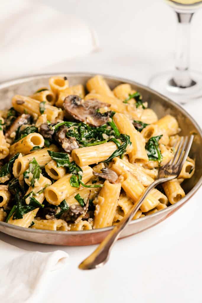 A plate of creamy Parmesan spinach mushroom pasta, with the penne coated in a light sauce, topped with sautéed mushrooms and fresh, chopped spinach, served on a metal pan with a fork on the side, all set on a white tablecloth with a glass of white wine in the background.