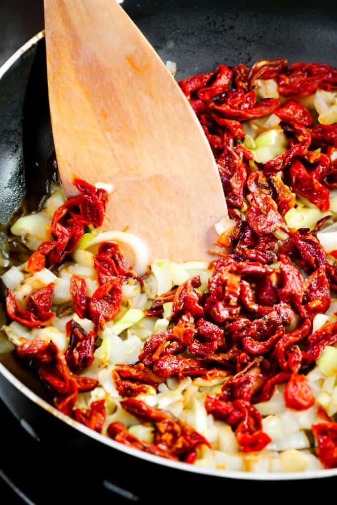 onions, garlic and sundried tomatoes cooking in a pan with a wooden spoon.