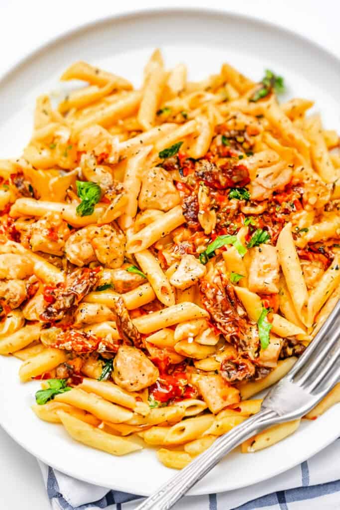 a white plate with Sun-Dried Tomato Pasta with a creamy sauce and chicken and a fork.