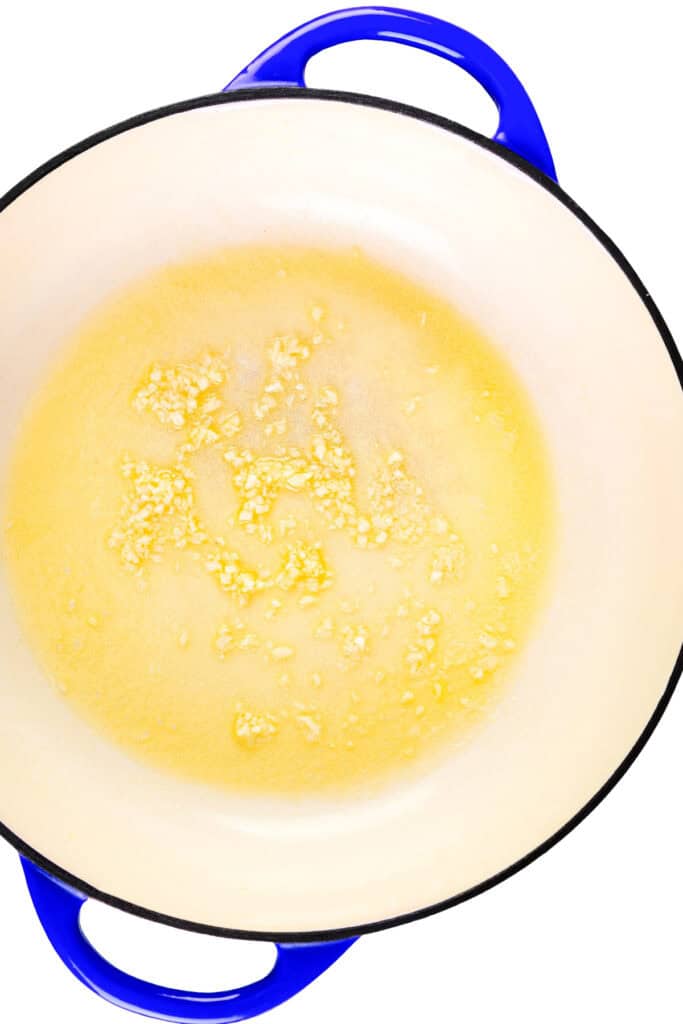 Chopped garlic sautéing in melted butter in a white enameled pan with blue handles, the perfect start for a flavorful Parmesan Lemon Orzo Pasta, viewed from above on a white background.