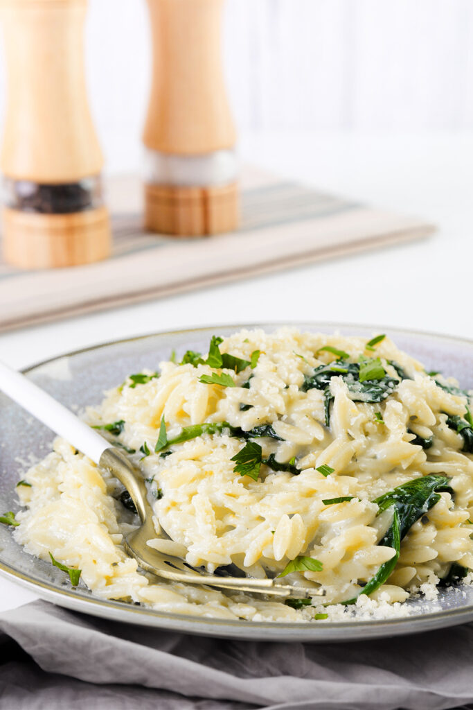 A plate of creamy Parmesan Lemon Orzo Pasta mixed with spinach and topped with grated cheese and fresh parsley, served with a spoon. Two wooden salt and pepper grinders are blurred in the background.