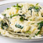 A plate of creamy Parmesan Lemon Orzo Pasta mixed with wilted spinach, topped with grated cheese and chopped fresh herbs, with a fork resting on the side.