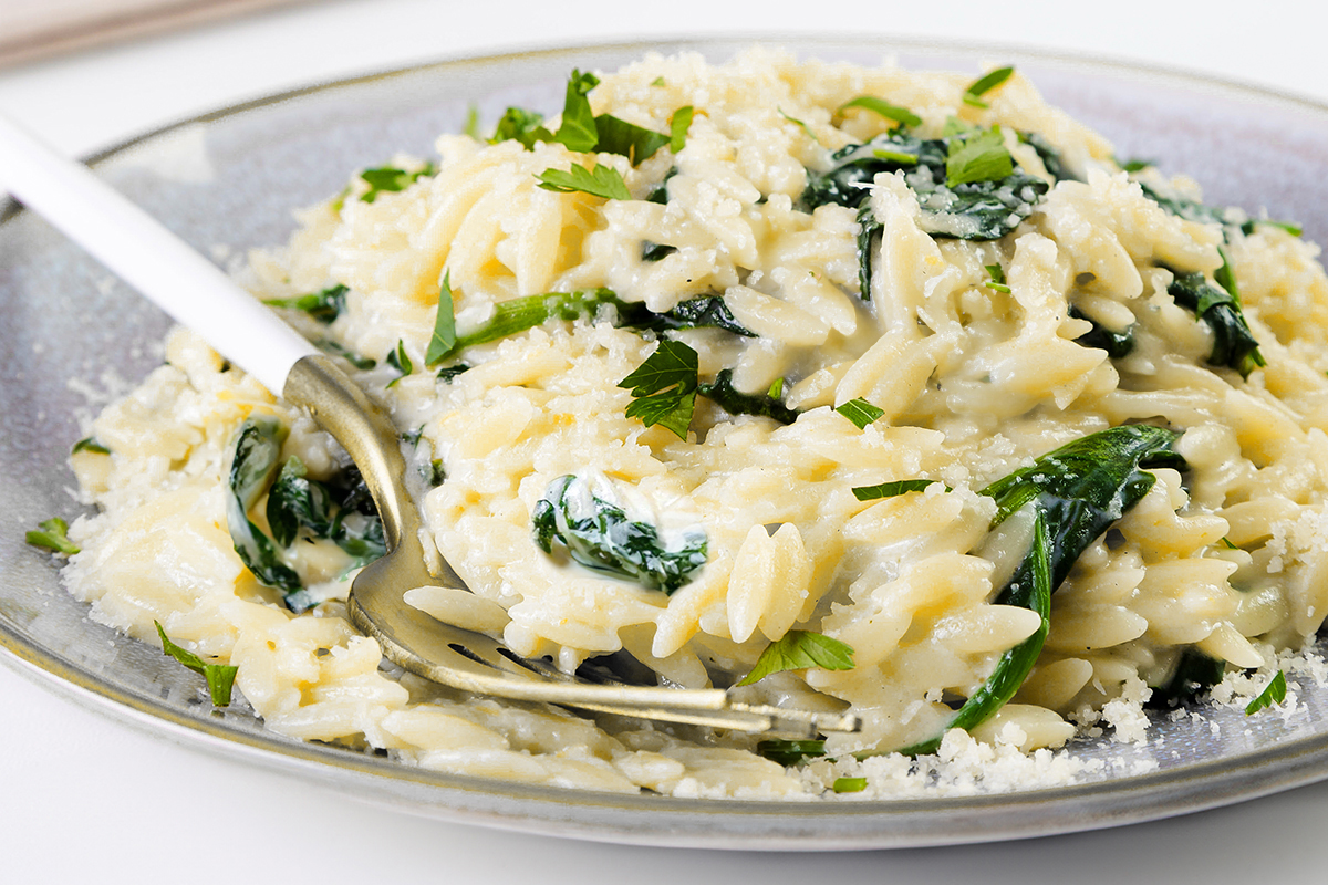 A plate of creamy Parmesan Lemon Orzo Pasta mixed with wilted spinach, topped with grated cheese and chopped fresh herbs, with a fork resting on the side.
