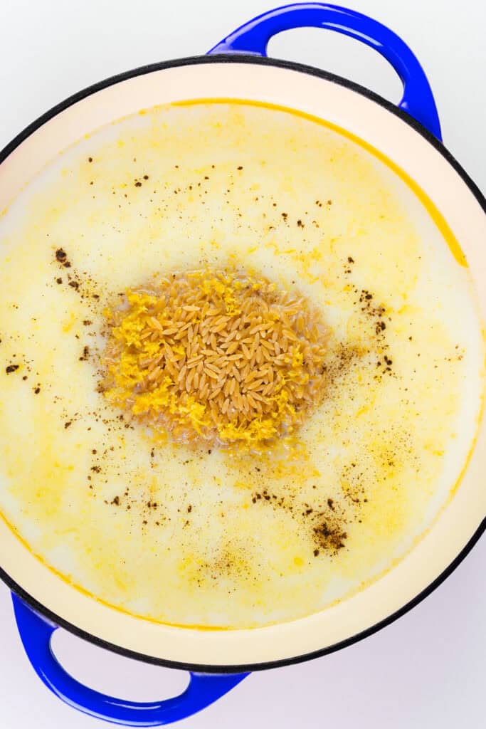 A blue pot filled with creamy white soup, topped with uncooked Parmesan Lemon Orzo Pasta and sprinkled with black pepper and yellow seasoning in the center.