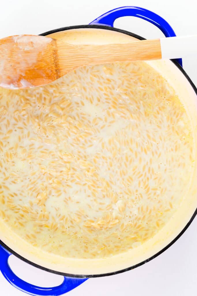 A blue-handled pot filled with creamy Parmesan Lemon Orzo Pasta simmers, with a wooden spoon resting on the edge of the pot.
