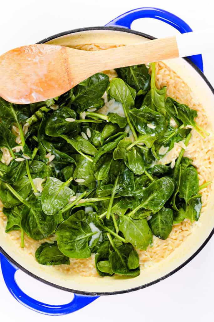 A blue pot filled with Parmesan Lemon Orzo Pasta in creamy sauce and fresh spinach leaves, with a wooden spoon resting on top.