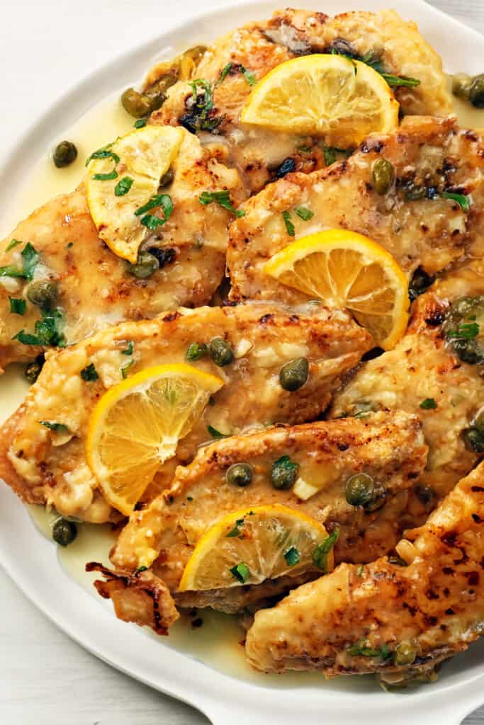 A platter full of fish piccata garnished with lemon slices and chopped herbs.