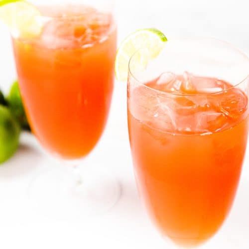 Two tall glasses filled with reddish-orange Bay Breeze Cocktails, each garnished with a lime wedge. Three whole limes rest nearby on a white surface, set against a soft white background.