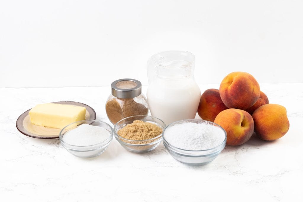 Butter on a plate, a jar of cinnamon, a jug of milk, four peaches, and bowls of white sugar, brown sugar, and flour are arranged on a white surface—everything you need to create the perfect peaches and cream dessert.