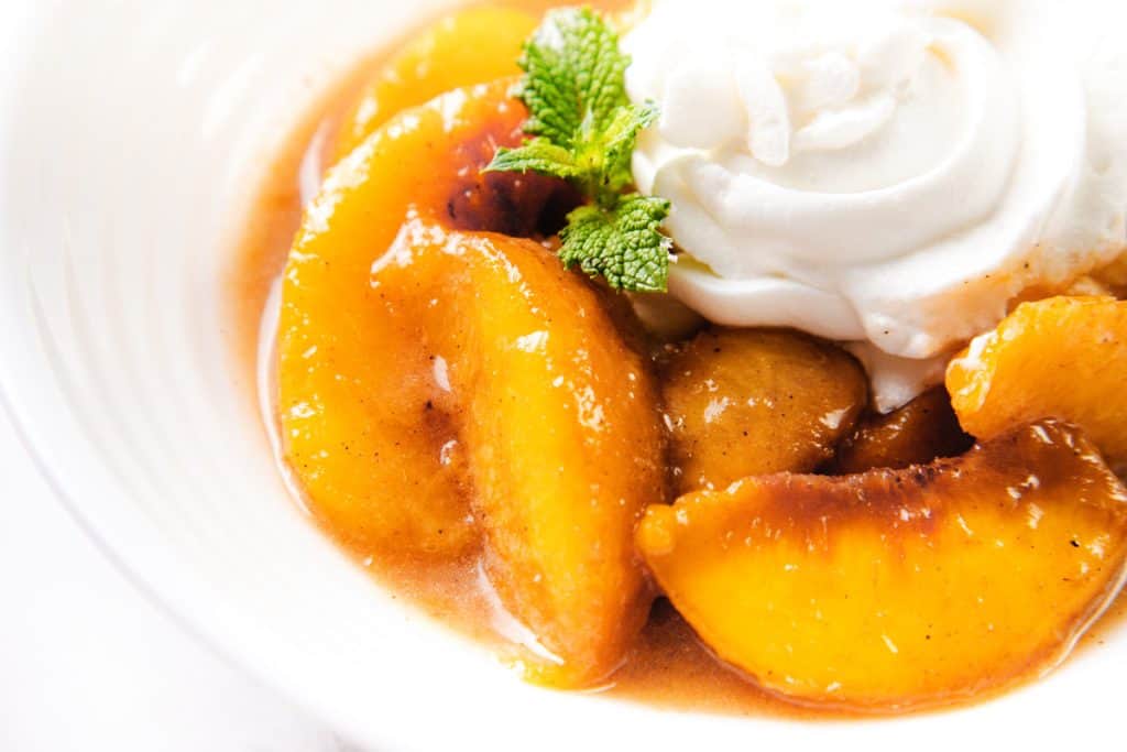 Peaches and cream come together in this delightful dessert: peach slices in syrup served in a white bowl, topped with whipped cream and garnished with a sprig of mint.