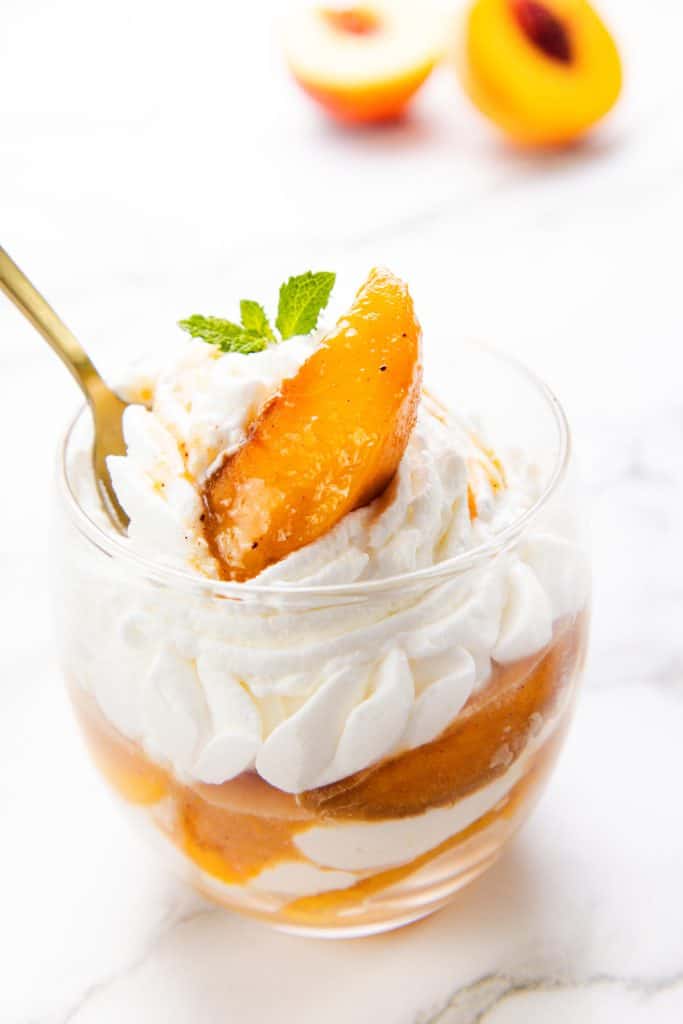 A glass cup filled with luscious whipped cream and juicy peach slices, garnished with a fresh mint leaf, captures the classic charm of Peaches and Cream. A gold spoon is inserted, while halved peaches are artfully blurred in the background.