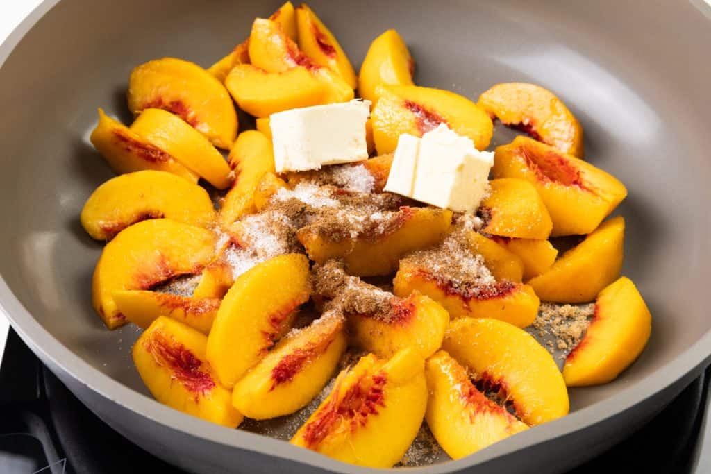 Sliced peaches in a pan topped with butter, sugar, and spices, ready to be cooked into a luscious peaches and cream-inspired dessert.
