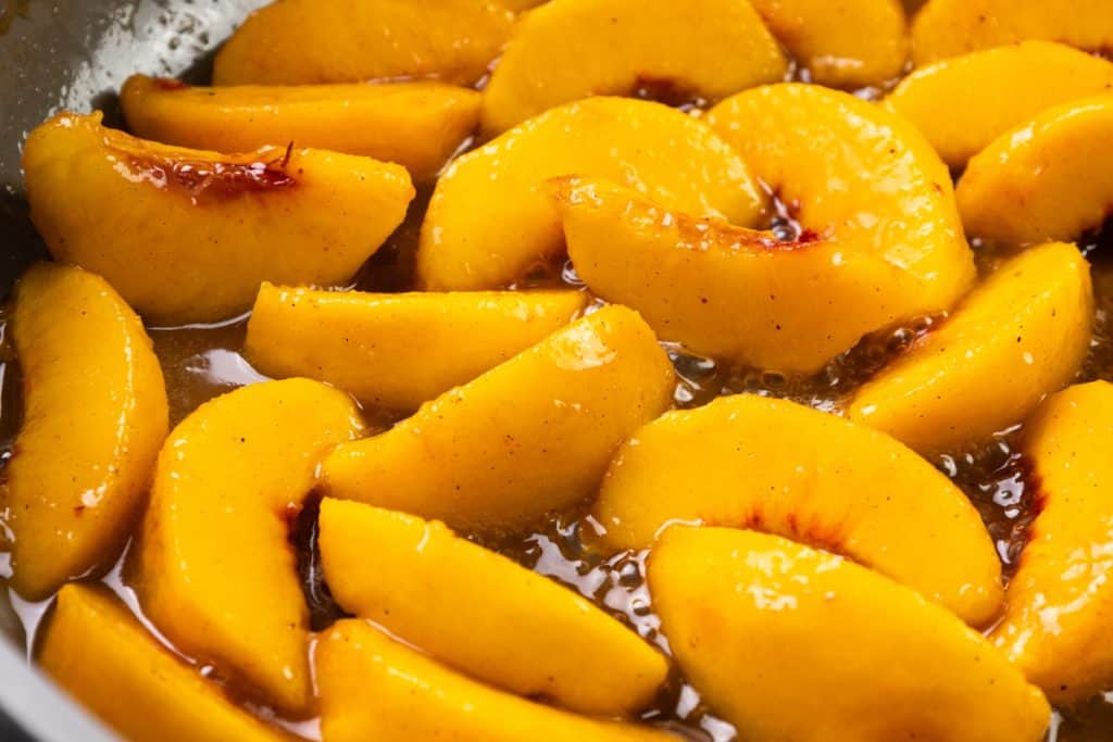 Sliced peaches coated in a glossy syrup with visible specks of spices, likely cinnamon, evoke the comforting flavors of classic Peaches and Cream as they glisten in a pan or bowl.