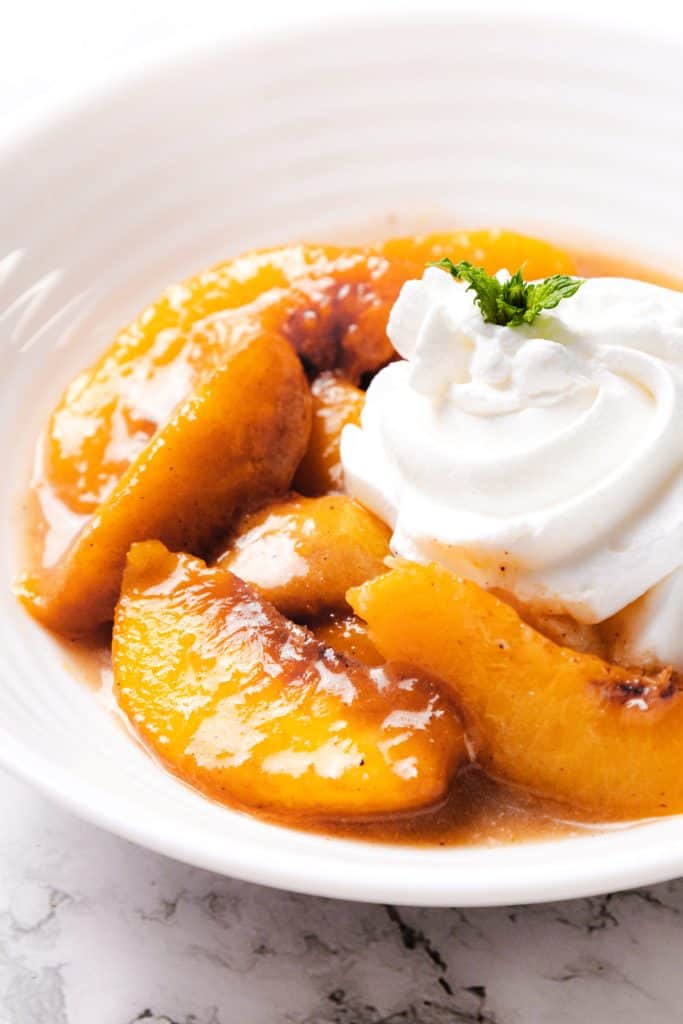 Sliced peaches in syrup served in a white bowl, topped with a swirl of whipped cream and a small mint garnish create the perfect Peaches and Cream dessert.