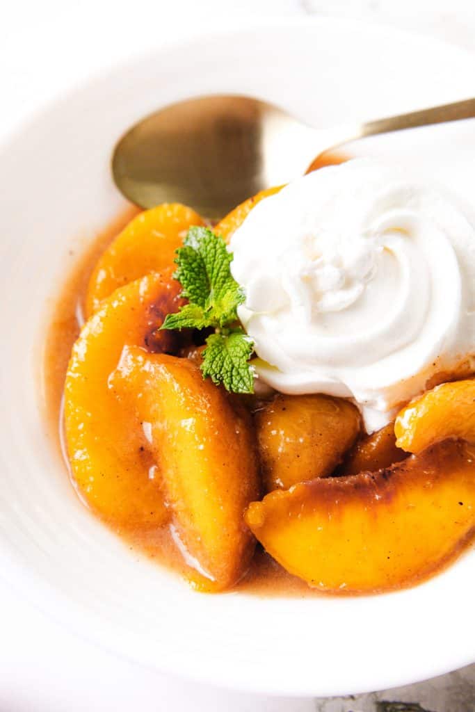 Sliced peaches in syrup topped with whipped cream and a sprig of mint, served in a white bowl with a spoon—this classic Peaches and Cream dessert is the perfect blend of fruity sweetness and silky richness.