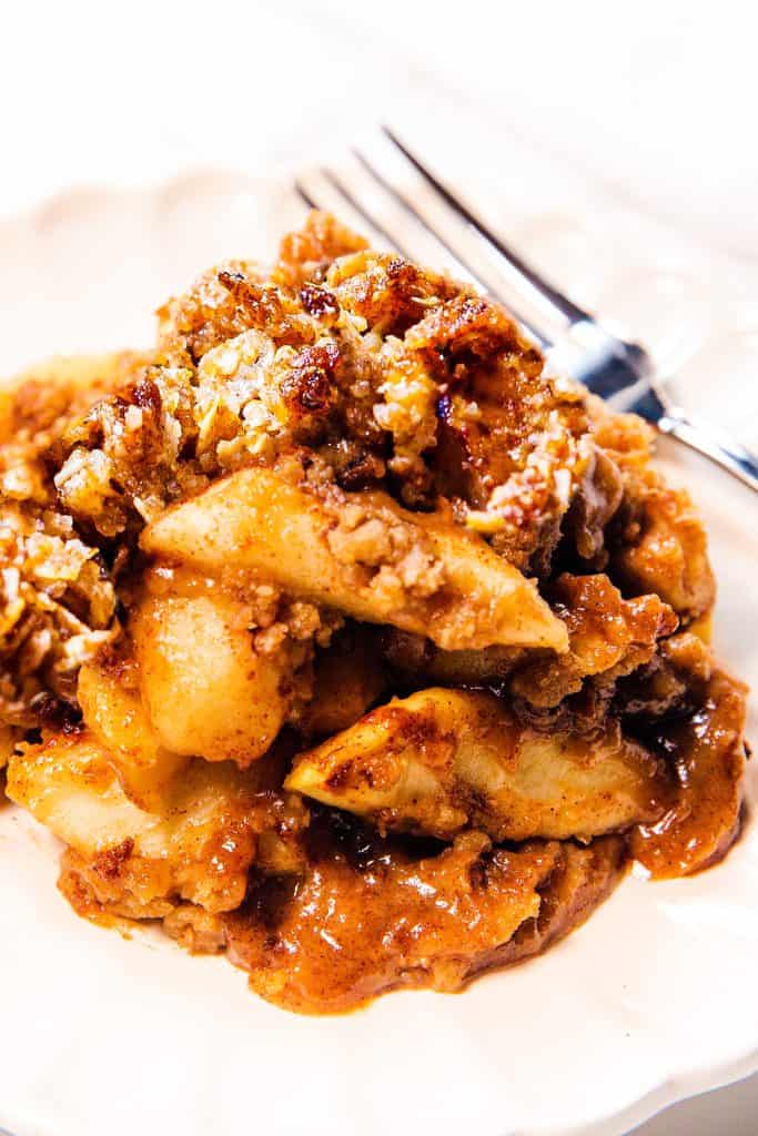 A close-up of a serving of Oatmeal Apple Crisp with tender baked apple slices and a crumbly oat topping on a white plate, with a fork in the background.