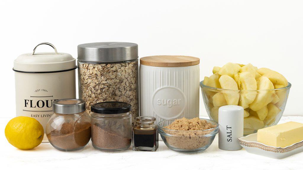 Baking ingredients for Oatmeal Apple Crisp on a white surface, including flour, oats, sugar, cinnamon, brown sugar, salt, vanilla, butter, a lemon, and a bowl of sliced apples in clear and labeled containers.