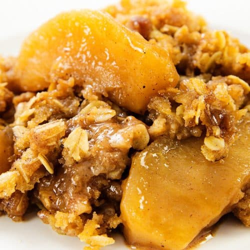 A close-up of Oatmeal Apple Crisp shows baked apple slices coated in cinnamon, sugar, and oats, with a golden-brown crumbly topping on a white plate.