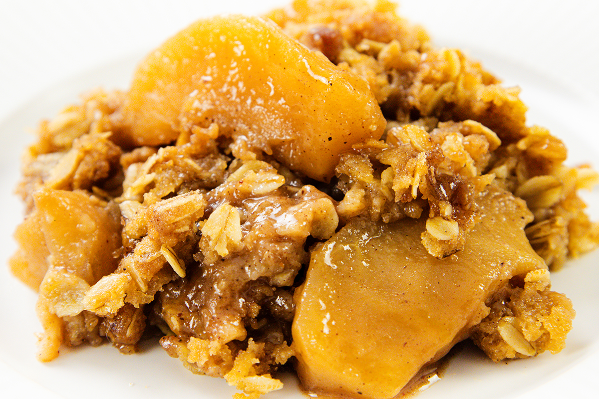 A close-up of Oatmeal Apple Crisp shows baked apple slices coated in cinnamon, sugar, and oats, with a golden-brown crumbly topping on a white plate.