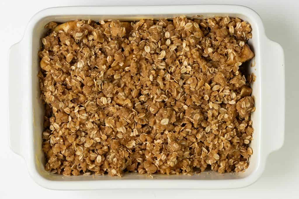 A white baking dish filled with golden brown Oatmeal Apple Crisp, topped with a crumbly oat and sugar mixture, seen from above on a white background.