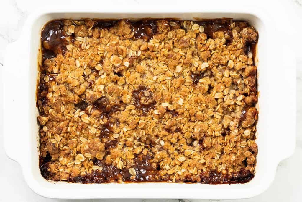 A baked Oatmeal Apple Crisp in a white rectangular dish, topped with a golden-brown oat crumble. The edges are slightly caramelized, and the oat topping appears crispy and textured.
