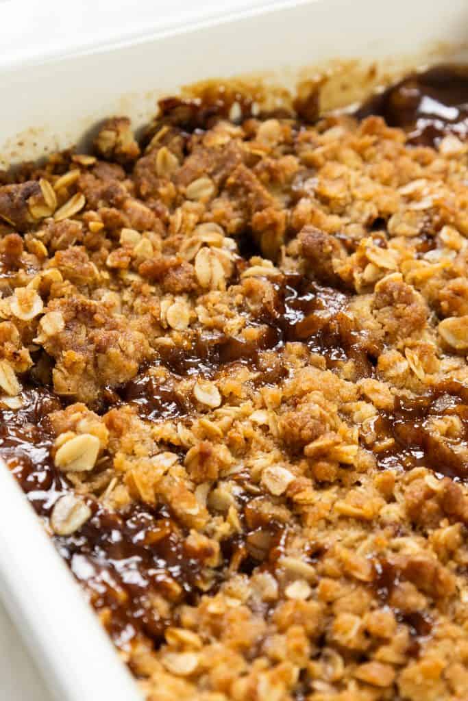 A close-up of an Oatmeal Apple Crisp in a white dish, showing a golden brown oat and crumb topping with bubbling caramelized sauce.