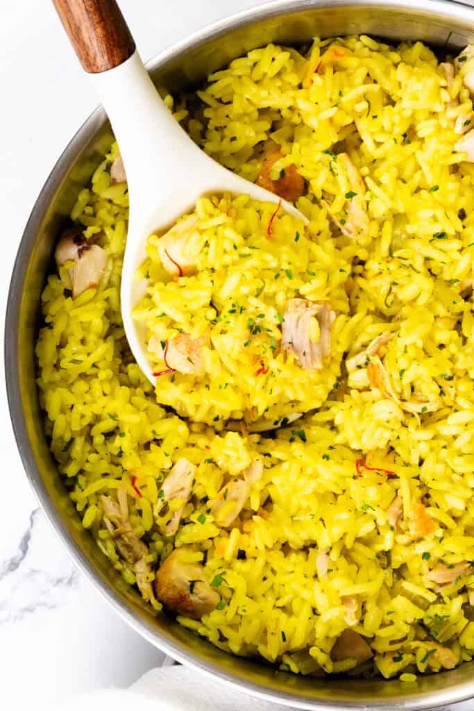 A pot of Chicken and Yellow Rice with small pieces of chicken is being served with a white spoon. The fluffy, seasoned rice is garnished with herbs, making the bright and inviting scene even more appetizing.