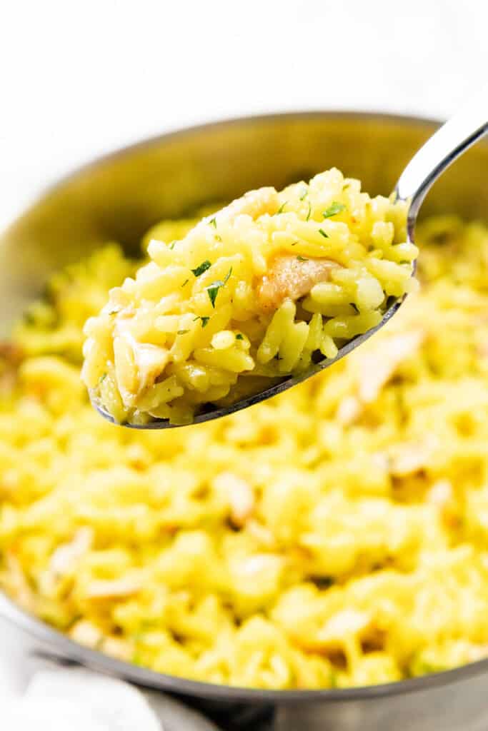 A close-up of a spoonful of cheesy Chicken and Yellow Rice with shredded chicken and herbs, held above a pan filled with more of the savory dish.