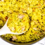 A close-up of a pot filled with Chicken and Yellow Rice, being stirred with a white spoon. The rice is garnished with herbs and has a vibrant, appetizing color.