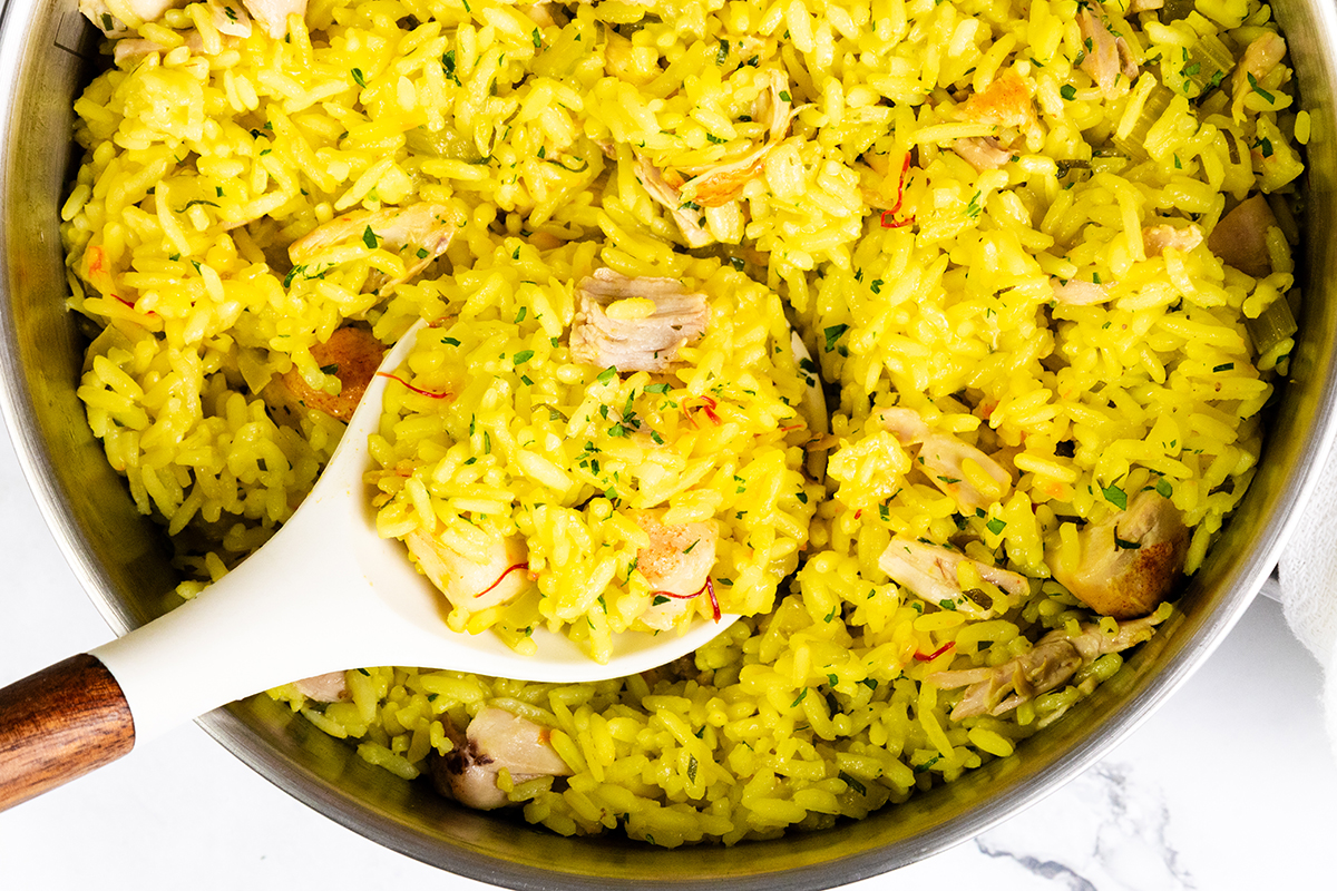 A close-up of a pot filled with Chicken and Yellow Rice, being stirred with a white spoon. The rice is garnished with herbs and has a vibrant, appetizing color.