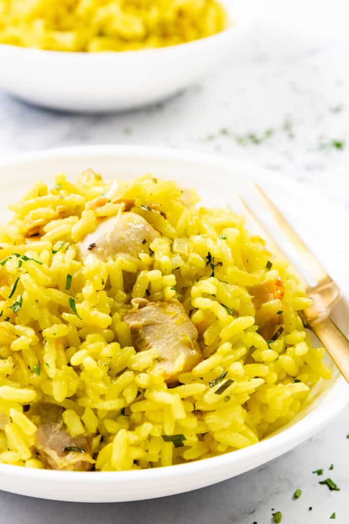 A close-up of a bowl of Chicken and Yellow Rice, garnished with herbs, beside a gold fork. Another bowl of rice is blurred in the background.