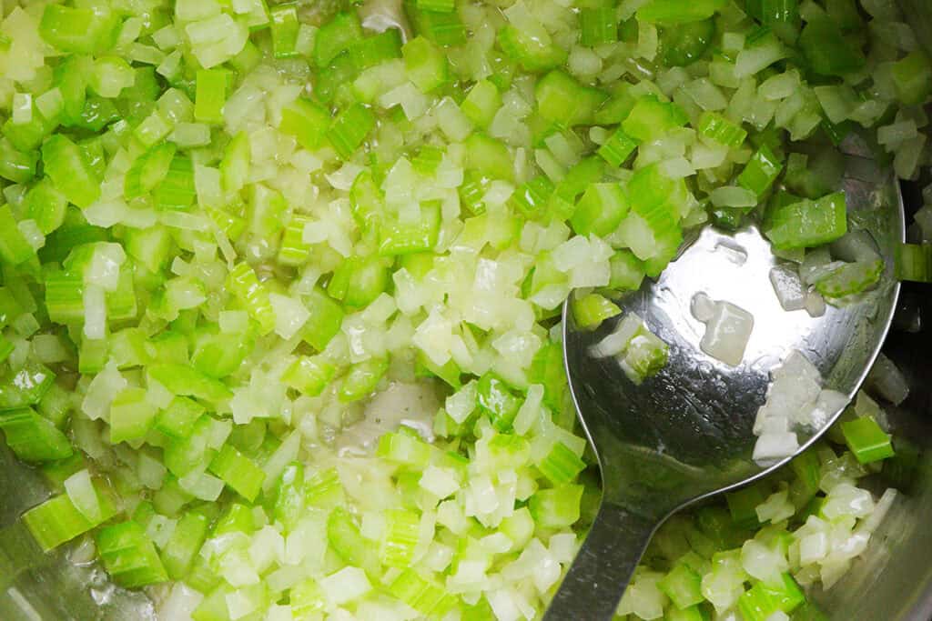 Close-up of chopped celery and onions being sautéed in a pan, with a metal spoon reflecting some vegetables—perfect for adding flavor to classic dishes like Chicken and Yellow Rice.