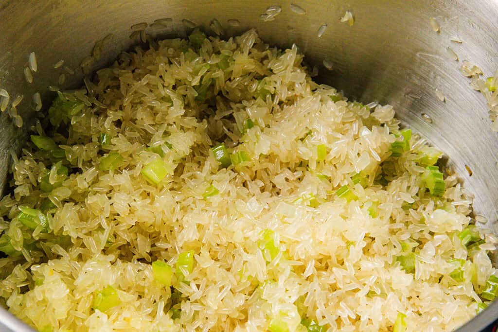 Uncooked rice mixed with chopped green celery pieces in a metal pot, perfect for preparing a delicious Chicken and Yellow Rice dish.