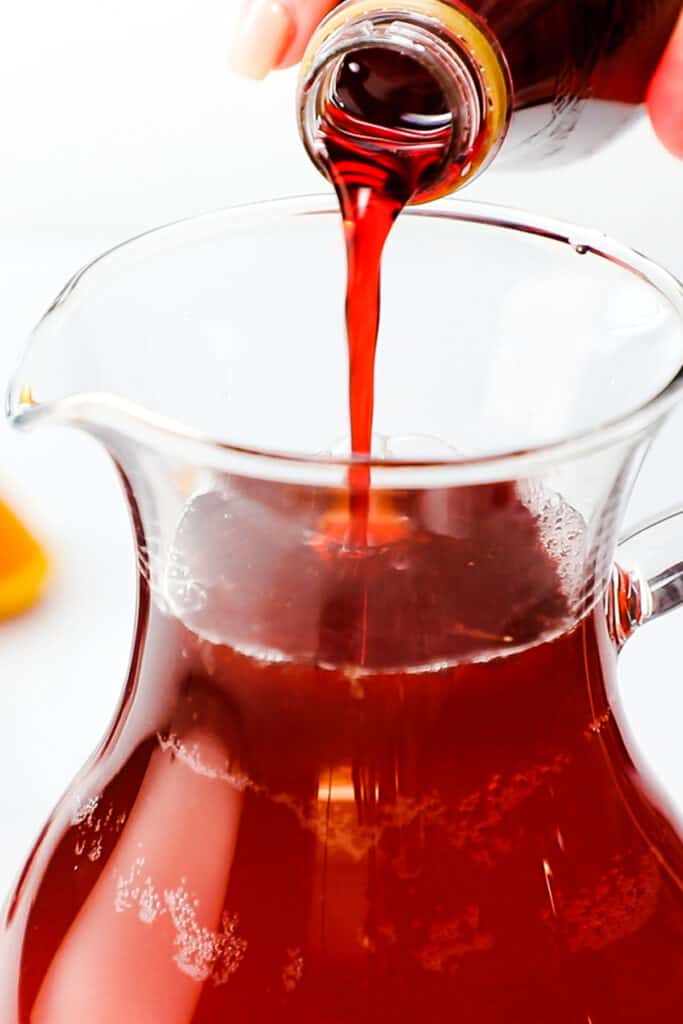 a woman pouring grenadine into the coktail in a pitcher