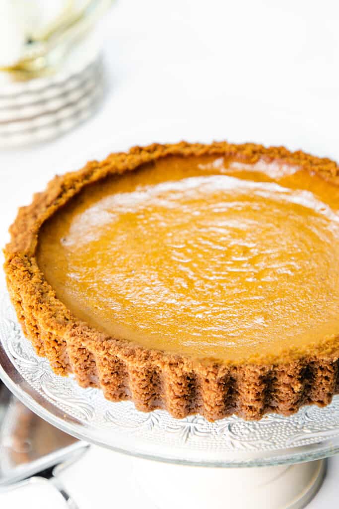 Pumpkin Pie with Graham Cracker Crust on a cake stand with a stack of plates in the background