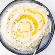 a close up photo taken from above of a bowl of Sour Cream Mashed Potatoes with melted butter and chives with a serving spoon