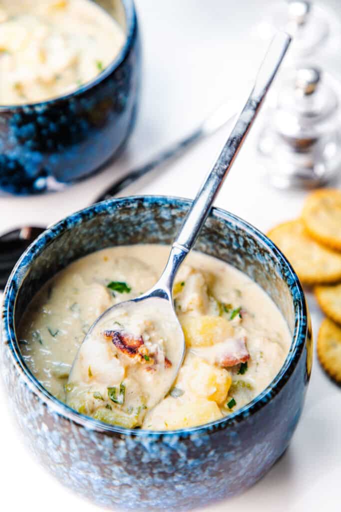 a bowl and spoon full of cream od crab soup with potatoes, cellery and crab meat with another bowl of soup behind it