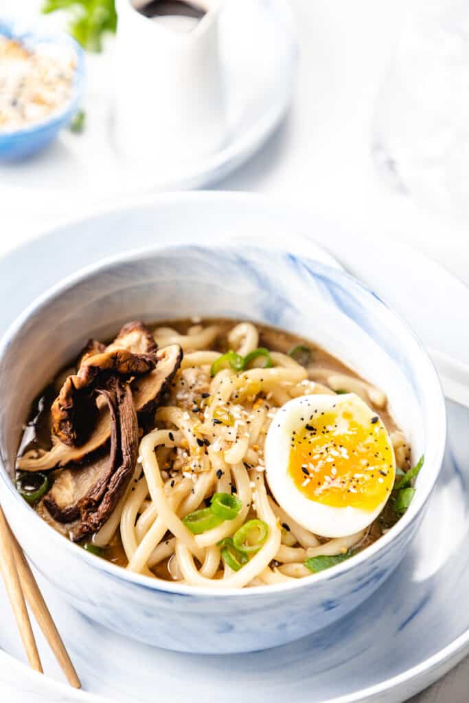 a bowl of udon soup topped with scallions, mushrooms, sesame seeds, and a soft-boiled