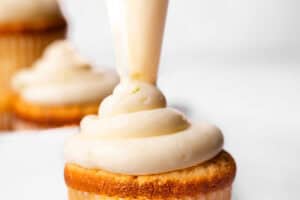 A close-up of a Key Lime Cupcake being frosted with creamy icing, showcasing the smooth and rich texture of the frosting with other frosted cupcakes blurred in the background.