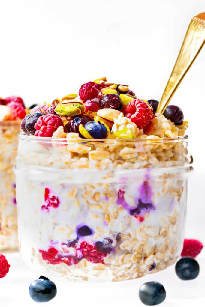 a jar of Overnight Oatmeal piled high with berries and chopped pistachios with a spoon in the jar.