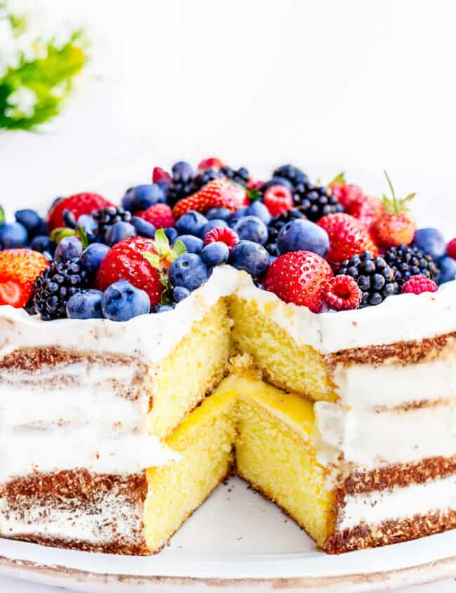 a lemon curd cake topped high with mixed berries with a slice cut out to reveal the lemon curd filling.