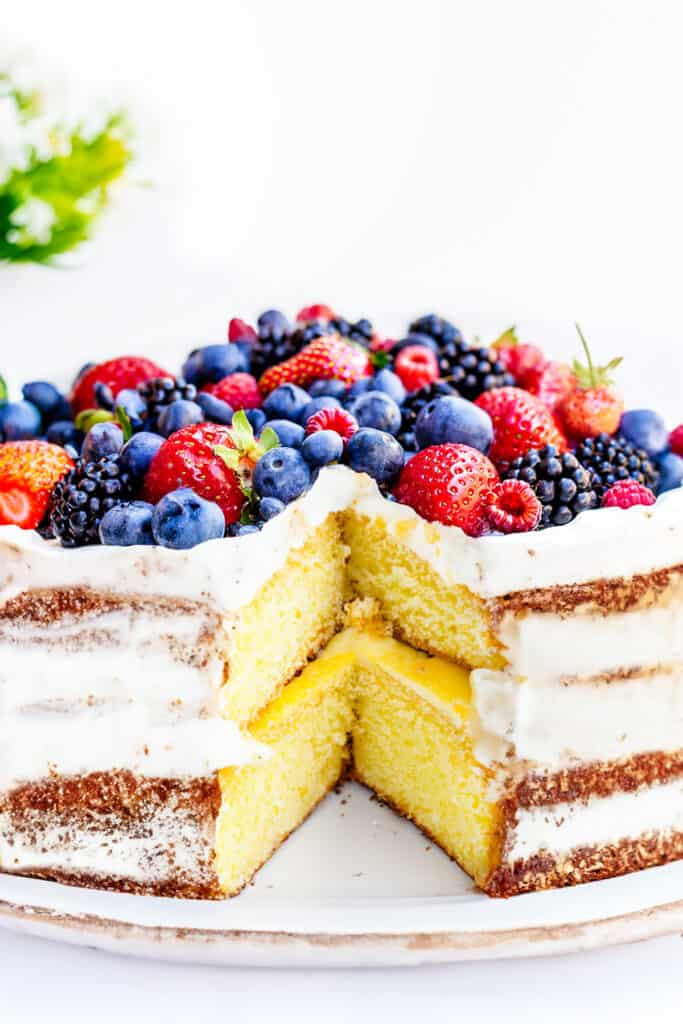 a lemon curd cake topped high with mixed berries with a slice cut out to reveal the lemon curd filling.